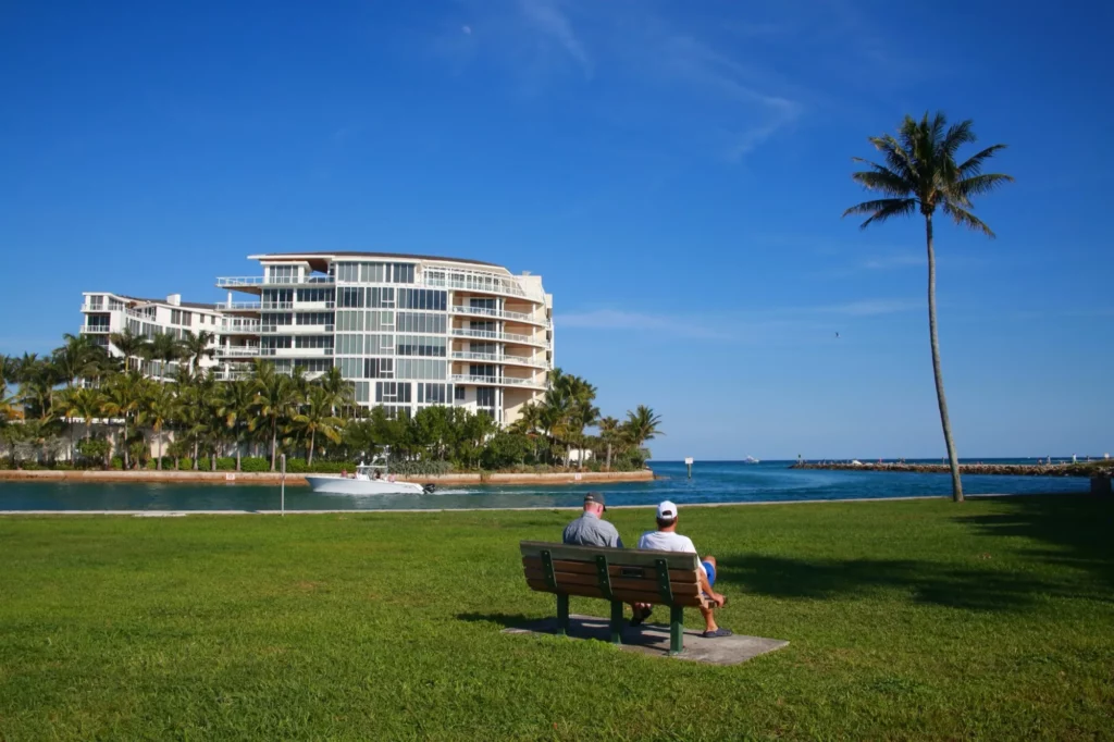 Boat passes in front of the Boca Beach Resort at the Inlet with two people sitting on a bench in the foreground in a clear sunny day